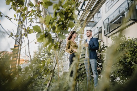 Zwei Personen stehen im Garten vor einem modernen Gebäude und unterhalten sich zwischen Pflanzen.