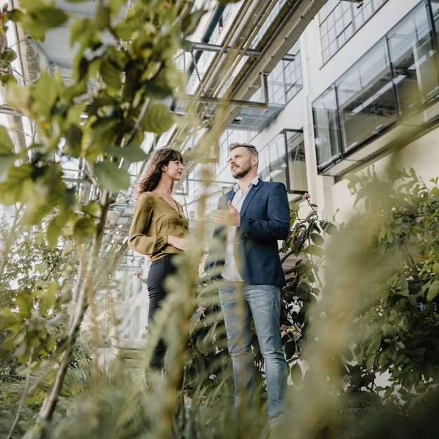 Zwei Personen stehen im Garten vor einem modernen Gebäude und unterhalten sich zwischen Pflanzen.