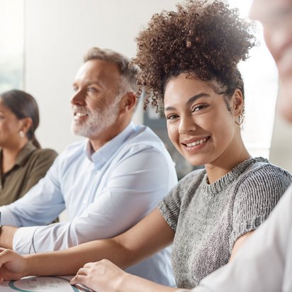 Lächelnde junge Frau mit Stift in der Hand sitzt mit anderen Personen in einem Seminarraum