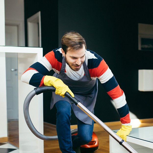 Eine Person in einem gestreiften Pullover und gelben Handschuhen reinigt mit einem Staubsauger den Boden eines modernen Wohnzimmers. Im Hintergrund sind Küchengeräte und Möbel zu sehen, die eine häusliche Umgebung darstellen.