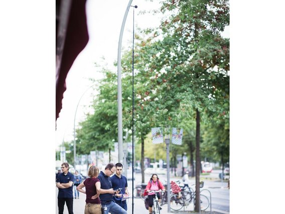 Eine kleine Gruppe von Personen steht auf einer Fußgängerzone vor Geschäften, während eine Frau mit Headset spricht und technisches Equipment für Aufnahmen aufgebaut ist.