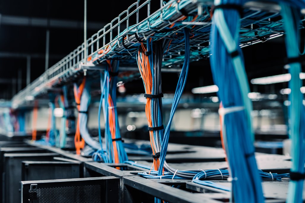 Nahaufnahme von Kabeln in einem Rechenzentrum, die ordentlich in Kabelkanälen verlegt sind. Die Kabel sind in verschiedenen Farben, hauptsächlich blau und orange, und verlaufen über Server-Racks, was eine organisierte und technologische Umgebung zeigt.