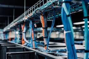 Nahaufnahme von Kabeln in einem Rechenzentrum, die ordentlich in Kabelkanälen verlegt sind. Die Kabel sind in verschiedenen Farben, hauptsächlich blau und orange, und verlaufen über Server-Racks, was eine organisierte und technologische Umgebung zeigt.