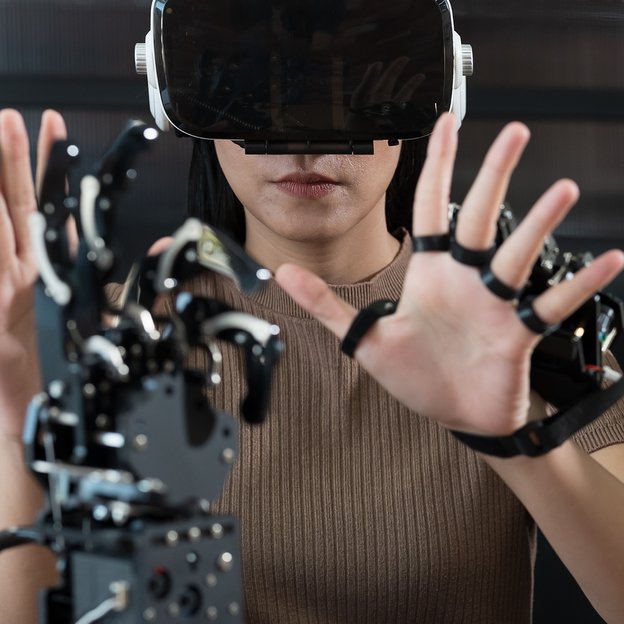 Eine Person mit VR- oder AR-Brille interagiert mit erhobenen Händen mit einer Roboterhand.