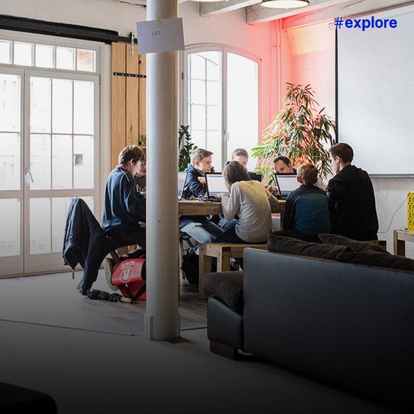 Ein moderner Arbeitsraum mit Holztüren, Pflanzen und einer Gruppe von Jugendlichen, die gemeinsam an einem Tisch an Computern arbeiten.