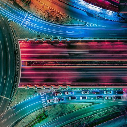 Luftaufnahme eines beleuchteten Autobahnknotenpunkts bei Nacht mit farbigen Lichtspuren von Fahrzeugen als Symbol für komplexe Mobilität und Verkehrsinfrastruktur