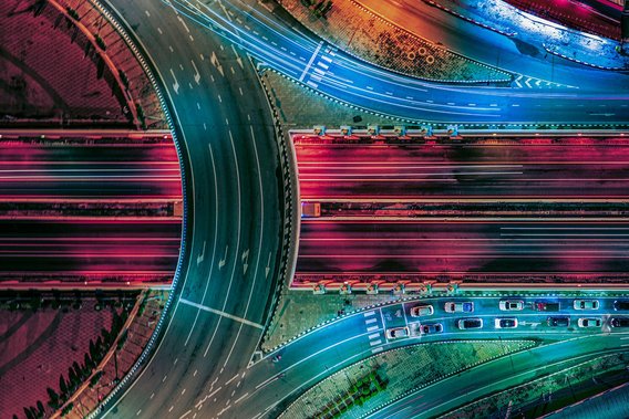 Urbane Mobilität im Wandel – Know-how Transfer für Fahrzeughersteller IFM Luftaufnahme eines beleuchteten Autobahnknotenpunkts bei Nacht mit farbigen Lichtspuren von Fahrzeugen als Symbol für komplexe Mobilität und Verkehrsinfrastruktur