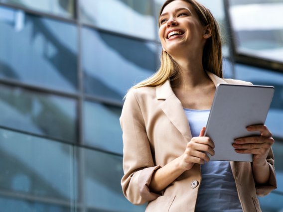 Junge Frau im Business-Outfit hält Unterlagen und lächelt vor einem modernen Bürogebäude.