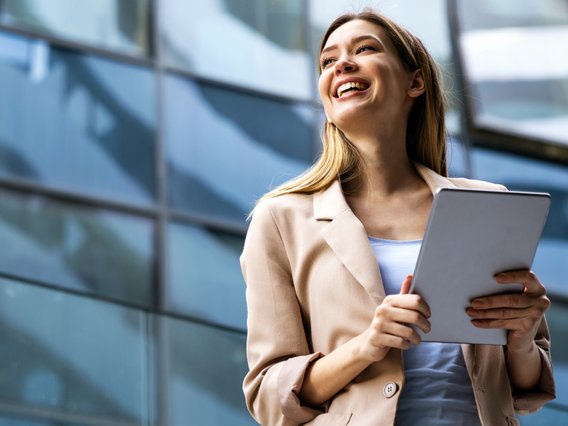 Junge Frau im Business-Outfit hält Unterlagen und lächelt vor einem modernen Bürogebäude.