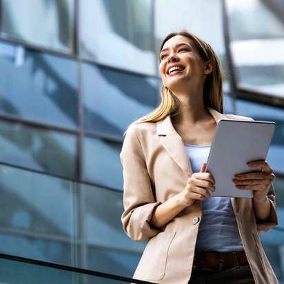 Junge Frau im Business-Outfit hält Unterlagen und lächelt vor einem modernen Bürogebäude.
