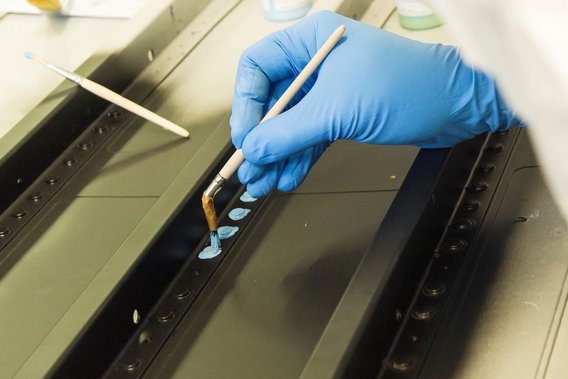 Eine Person mit blauen Handschuhen trägt mit einem Pinsel eine blaue Substanz auf ein Metallstück auf. Die Szene zeigt präzise Arbeit im Oberflächenschutz der Luftfahrtindustrie.