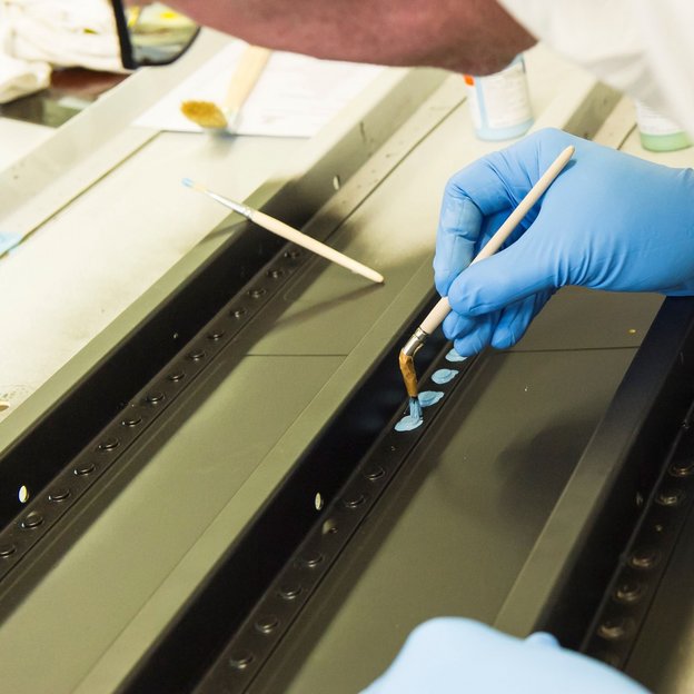 Eine Person mit blauen Handschuhen trägt mit einem Pinsel eine blaue Substanz auf ein Metallstück auf. Die Szene zeigt präzise Arbeit im Oberflächenschutz der Luftfahrtindustrie.