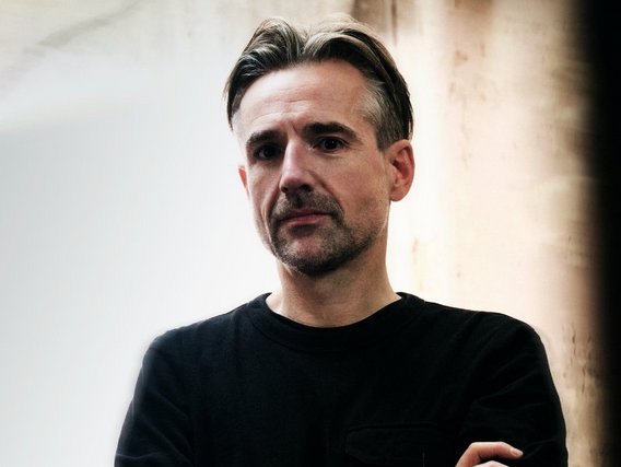 Bertolt Meyer in einem schwarzen T-Shirt steht vor einer hellen Wand und blickt nachdenklich.