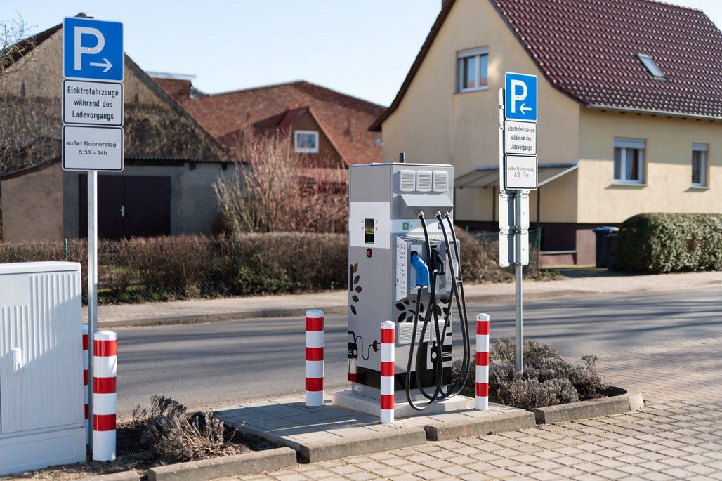 Öffentliche Elektroladesäule in einer Wohngegend mit zwei blauen Parkschildern für Elektrofahrzeuge. Die Ladestation mit mehreren Anschlüssen steht zwischen rot-weißen Absperrpfosten auf dem Gehweg vor Einfamilienhäusern.