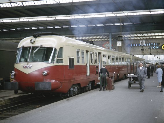Historischer rot-weißer Triebwagen mit ÖBB-Logo steht in einer überdachten Bahnhofshalle, Reisende in 1960er-Jahre-Kleidung auf dem Bahnsteig.