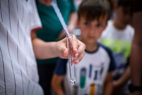 Eine Nahaufnahme eines wissenschaftlichen Experiments. Eine Person hält ein Reagenzglas oder eine Pipette, während Kinder im Hintergrund interessiert zuschauen.