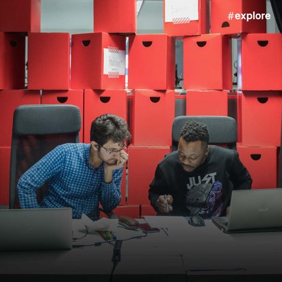 Zwei Personen arbeiten an Laptops an einem Tisch mit roten Aufbewahrungsboxen im Hintergrund.