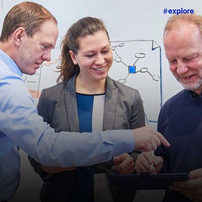 Drei Personen arbeiten zusammen an einem Whiteboard mit Skizzen und der Überschrift "IDEAS". Die Person links zeigt auf ein Tablet, während alle drei konzentriert auf das Gerät schauen.