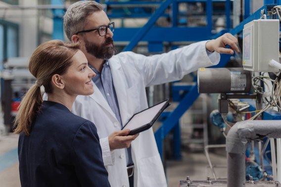 Ein technischer Experte erklärt einer Kollegin in einer Industrieanlage die Mess- und Kontrollsysteme. Beide stehen vor einem Schaltschrank, im Hintergrund sind Rohre und metallische Strukturen sichtbar.