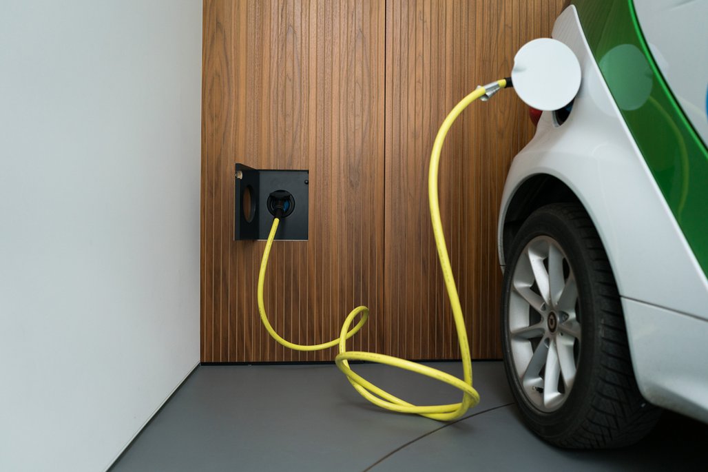 Eine Ladestation für Elektrofahrzeuge mit gelbem Kabel, das an ein weißes Auto angeschlossen ist. Die Ladestation ist in eine holzverkleidete Wand eingelassen.