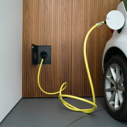 Eine Ladestation für Elektrofahrzeuge mit gelbem Kabel, das an ein weißes Auto angeschlossen ist. Die Ladestation ist in eine holzverkleidete Wand eingelassen.