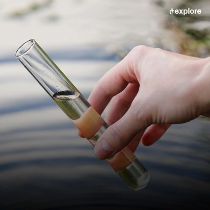Hand hält ein Reagenzglas mit klarer Flüssigkeit gegen verschwommenen Hintergrund mit Wasser und Steinen.