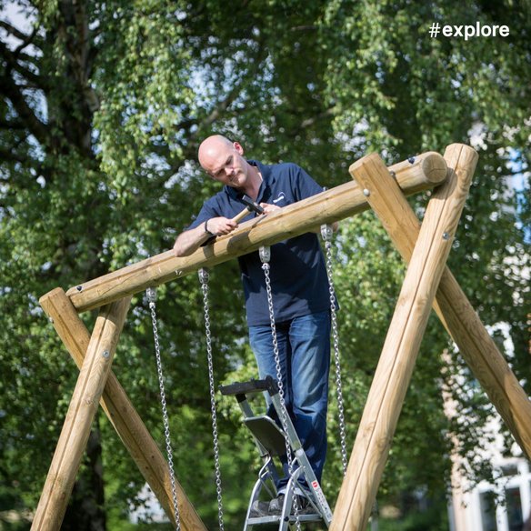 Der Arbeiter steht auf einer Leiter und arbeitet an der Montage einer Holzkonstruktion mit Ketten. Das Spielgerät befindet sich in einem Park mit grünen Bäumen im Hintergrund.