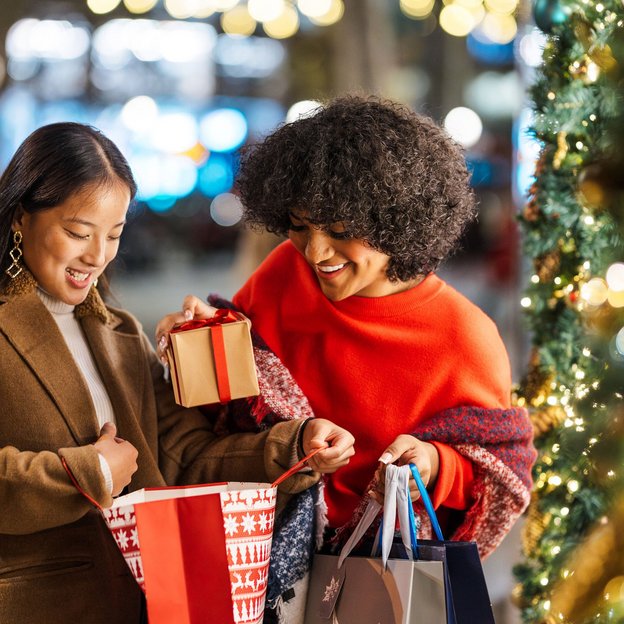 Zwei Frauen, die vor einem festlich beleuchteten Baum Geschenke aus Taschen nehmen und lächeln.