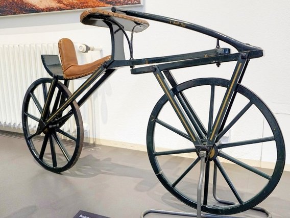 Ein historisches Fahrrad in einem Museum, mit ungewöhnlichem Design und ledersitzem Sattel. Schwarze Metallrahmen und große Räder zeigen die frühe Ingenieurskunst.