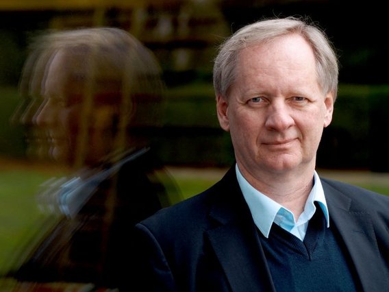 Porträt von Ulrich Eber in dunklem Anzug und hellblauem Hemd. Im Hintergrund ist eine verschwommene Spiegelung seines Gesichts 