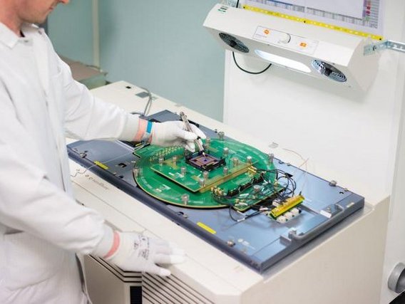 Ein Techniker in Schutzkleidung und Handschuhen bearbeitet eine grüne Elektronikplatine an einem Arbeitsplatz. Über dem Arbeitsbereich befindet sich eine Absaugvorrichtung für Dämpfe.