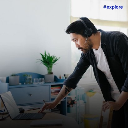 Ein Mann mit Kopfhörern und dunklem Pullover arbeitet konzentriert an seinem Laptop in einer hellen, modernen Küche. Er steht am Tresen und ist von verschiedenen Küchengeräten und einer Pflanze umgeben.