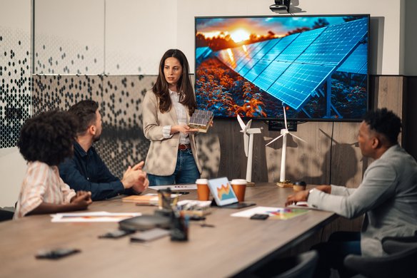 Frau präsentiert in Meeting über erneuerbare Energien mit Modellen von Windrädern und Solarenergie auf dem Bildschirm.