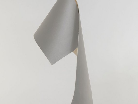 Eine Hand hält ein graues, flexibles Material vor neutralem Hintergrund, das sich elegant faltet und biegt. Das Material zeigt eine matte Oberfläche mit natürlicher Textur und könnte nachhaltiges Leder oder biobasiertes Textilmaterial darstellen.