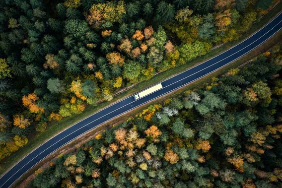 Luftaufnahme eines Lastwagens, der auf einer kurvenreichen Straße durch einen herbstlich gefärbten Wald fährt.