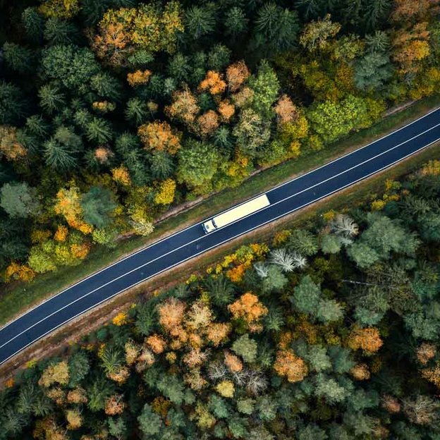 Luftaufnahme eines Lastwagens, der auf einer kurvenreichen Straße durch einen herbstlich gefärbten Wald fährt.