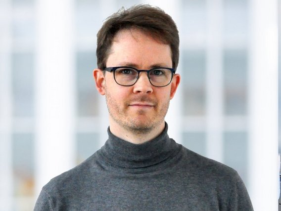 Porträtaufnahme von Phillip Denger mit schwarzer Brille und grauem Rollkragenpullover vor einem hellen, unscharfen Büro-Hintergrund.