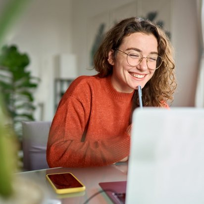  Junge Frau mit Brille und rotem Pullover sitzt lächelnd vor einem Laptop am Schreibtisch