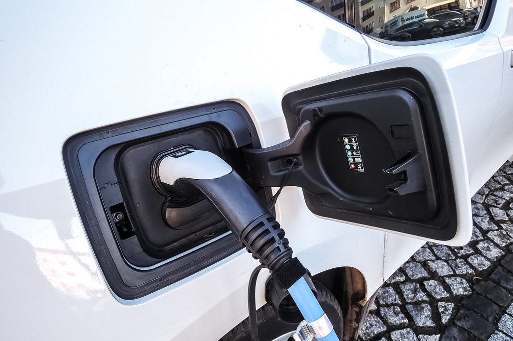 Ein weißes Elektroauto mit geöffneter Ladeklappe, in der ein schwarzer Ladestecker mit blauem Akzent steckt. Der Ladeport ist an der Seite des Fahrzeugs angebracht und das Auto steht auf einem gepflasterten Untergrund.