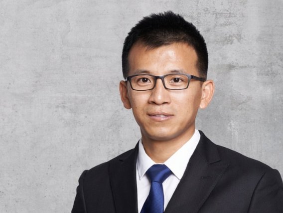 Hao Zhong in einem dunklen Anzug mit blauer Krawatte und Brille vor einem grauen Hintergrund, vermutlich ein Geschäfts- oder Bewerbungsfoto.
