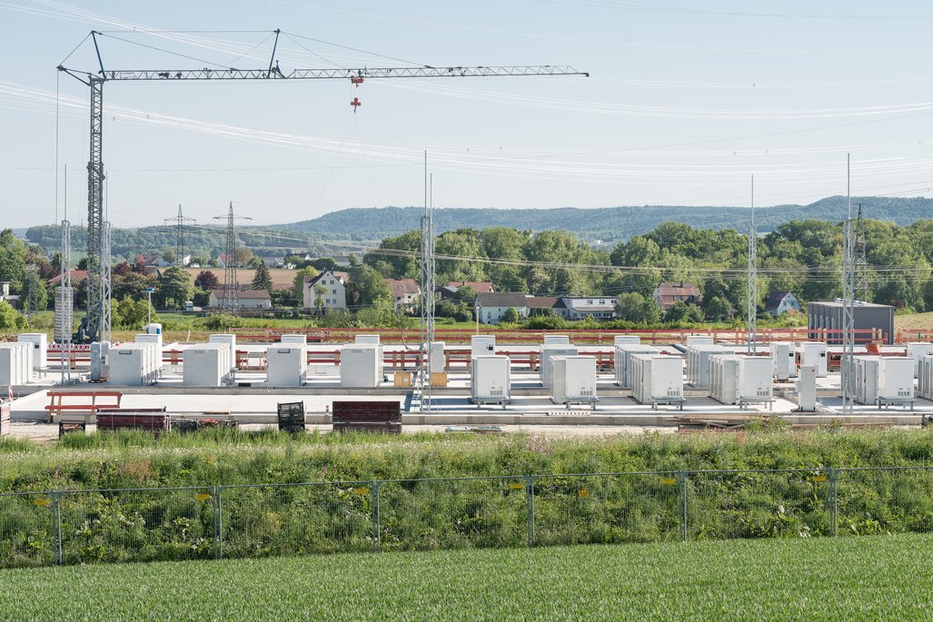 A construction site featuring several white container buildings on a concrete foundation. A large crane towers over the facility, with power lines and a rural landscape of houses and hills visible in the background.