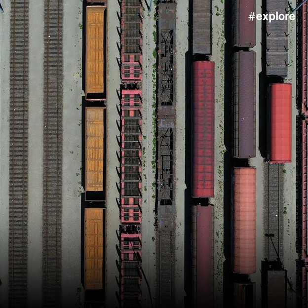 Luftaufnahme eines großen Güterbahnhofs mit parallelen Gleisen und bunten Güterwaggons in verschiedenen Farben (rot, orange, braun).