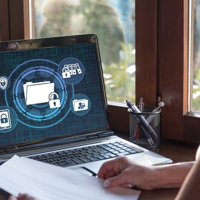 Laptop mit Datenschutzsymbolen auf dem Bildschirm und Person mit Dokument am Schreibtisch.