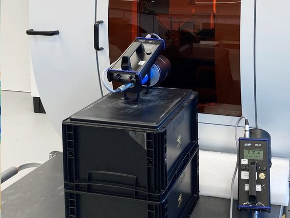 Industrielle Messeinrichtung mit blauem Scanner-Gerät auf schwarzen Transportboxen und einem digitalen Messgerät, positioniert vor einer Glasfläche in einem technischen Arbeitsbereich.
