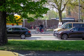 Straßenszene mit Radfahrer auf rotem Leihrad zwischen parkenden Autos und einem Fahrradparkplatz.
