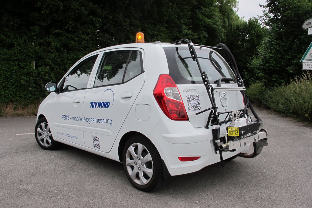 Ein weißes Auto mit dem Logo von TÜV NORD und der Aufschrift "PEMS - mobile Abgasmessung" steht auf einem Parkplatz. Auf der Rückseite des Fahrzeugs ist ein komplexes Messgerät montiert, das für die mobile Abgasmessung verwendet wird. Das Auto hat ein gelbes Warnlicht auf dem Dach. Auf der Seite des Fahrzeugs und am Heck sind QR-Codes angebracht. Im Hintergrund sind grüne Büsche und Bäume zu sehen.