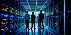 Vier Personen stehen in einem beleuchteten Gang eines modernen Rechenzentrums, umgeben von Server-Racks, die mit blauen und gelben Lichtern leuchten. Die Umgebung vermittelt eine hochtechnologische und futuristische Atmosphäre.