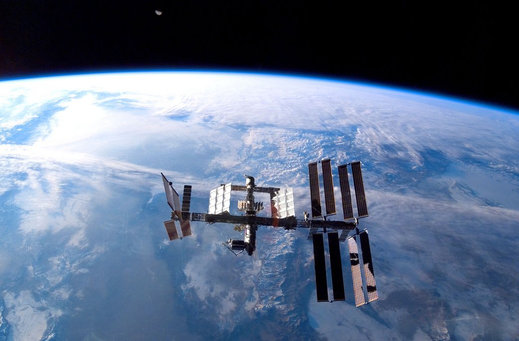 Die Internationale Raumstation (ISS) schwebt über der Erde mit ausgebreiteten Solarpaneelen, dahinter ist die gekrümmte blaue Erdatmosphäre und Wolkenformationen sichtbar.