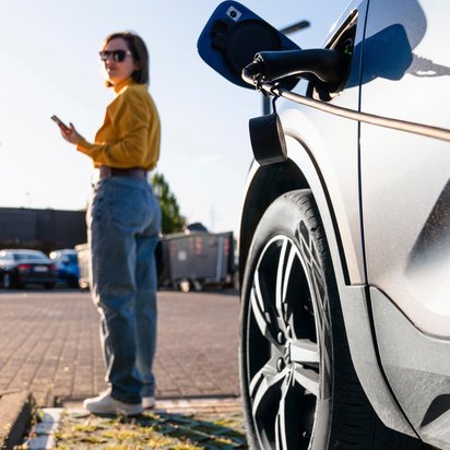Frau steht neben einem Elektroauto an einer Ladestation und nutzt ihr Smartphone.