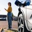 Frau steht neben einem Elektroauto an einer Ladestation und nutzt ihr Smartphone.